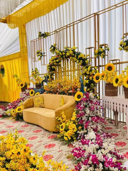 The main seating area for a sunflower-themed Haldi. A mustard yellow sofa is surrounded by an explosion of sunflowers and other florals against a modern gold-framed backdrop.