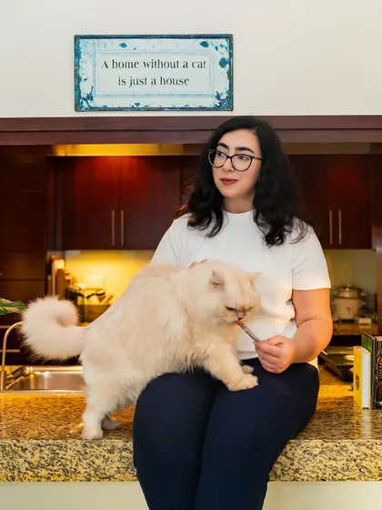 The sign says it all: "A home without a cat is just a house." This lifestyle shot captures a quiet moment in the kitchen, a woman sharing a treat with her beautiful Persian cat.