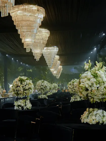 A dramatic, all-black seating arrangement for a modern cocktail event. The space is punctuated by large white floral centerpieces and magnificent tiered crystal chandeliers.