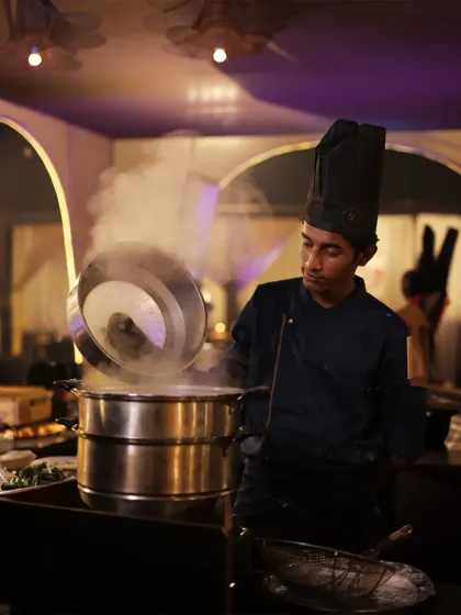 A chef carefully lifts the lid of a steamer, revealing perfectly cooked dumplings. The rising steam creates a sense of anticipation and highlights the freshness of our food.