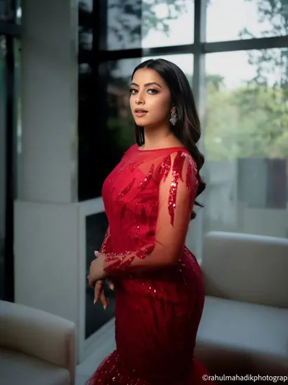 A striking portrait of a woman in a glamorous red gown. The indoor setting with large windows provides soft, natural light that highlights her elegance and the dress's sparkling details.