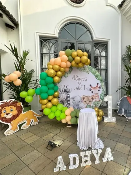 A 'Wild One' jungle safari theme perfect for an outdoor first birthday. This decoration features a custom backdrop with cute safari animals, an organic balloon garland in green and gold, and character cutouts.