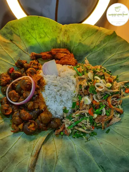 A special meal served on a lotus leaf, featuring spicy chicken salad, river snail curry (tharoi thongba), sticky rice, and a chicken drumstick.