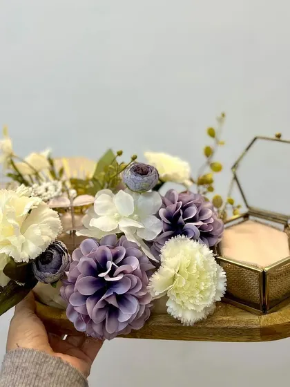 An engagement ring platter we designed, featuring a beautiful arrangement of purple and white flowers on a wooden tray. The hexagonal glass box adds a modern touch.