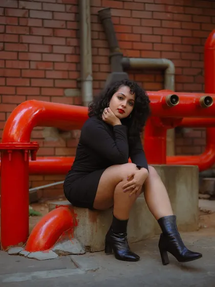 A full-body shot of a model in a black dress against a contrasting red and brick background. The composition is strong and highlights her entire look.