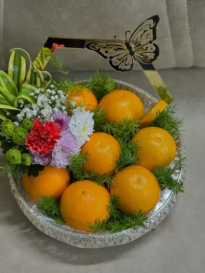 A simple yet elegant fruit tray design. Fresh oranges are nestled in a bed of green foliage within a silver platter, accented with a modern, gold-rimmed butterfly handle for a touch of contemporary style.