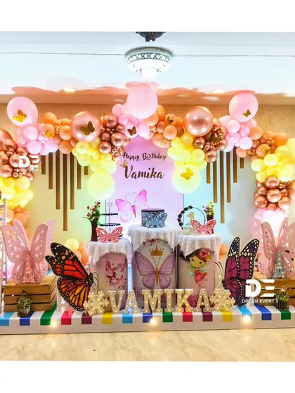 A warm and inviting butterfly-themed party for Vamika. This setup uses a beautiful color palette of rose gold, pink, and yellow balloons, with large butterfly wings and a striped floor mat to complete the look.
