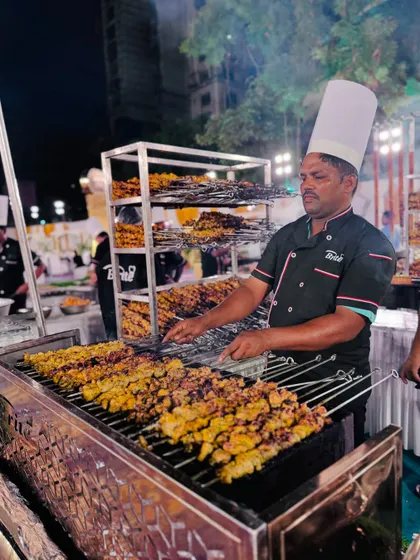 A chef at the live BBQ station, grilling skewers of Chicken Zafrani Tikka Barra. The multi-level racks behind him hold dozens of freshly cooked kebabs.