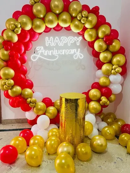 A classic red, white, and gold balloon ring for an anniversary. This setup includes a "Happy Anniversary" neon sign and a gold pedestal for a cake or gifts.