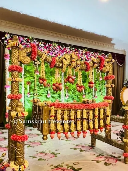 A traditional wooden cradle decorated with garlands of red and yellow flowers. The green backdrop and floral carpet create a vibrant and natural setting for the baby's naming ceremony.