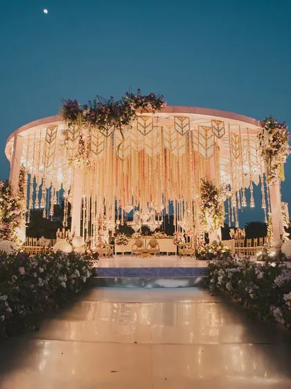 An open-air circular mandap under the evening sky, adorned with cascading floral strings and intricate latticework. This design creates a 360-degree experience of grandeur and romance.