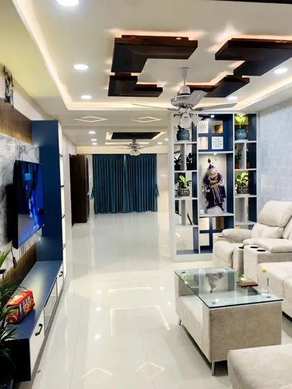 A spacious and luxurious living room we designed. The room features a large TV unit, a sectional sofa, and a false ceiling with intricate lighting, all tied together with a cool blue and white color scheme.
