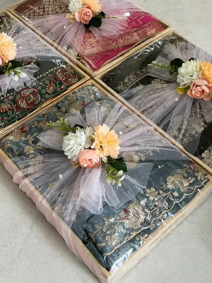 A collection of trousseau gifts packed in light pinewood trays. The pastel tulle wrapping and floral details add a soft, romantic touch to the embroidered outfits.