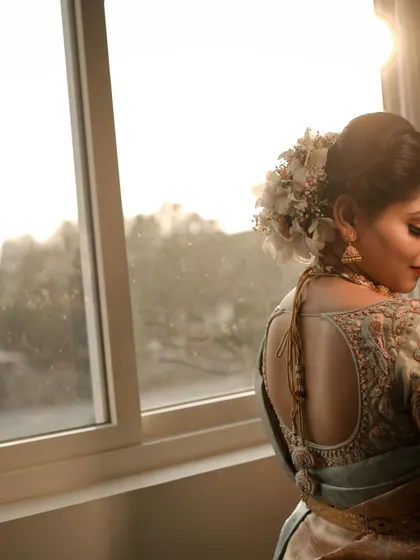A profile shot of a bride by a window, showcasing the elegant design of her blouse back and her floral hair arrangement.