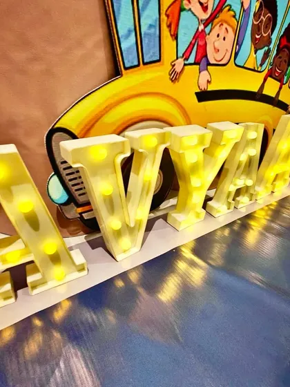 Personalized light-up marquee letters spelling 'AVYAAN' in front of a school bus cutout, part of the car-themed decor.