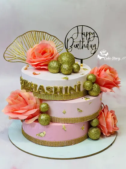 An elegant three-tier cake in shades of pink and gold. This crisscross design is adorned with sparkling gold balls and soft peach roses, perfect for a grand birthday celebration.