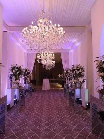 A grand entrance corridor designed for a reception at The Oberoi, Gurgaon. The walkway is lined with large floral arrangements on mirrored pedestals, illuminated by purple uplighting and magnificent chandeliers, setting a tone of opulence.