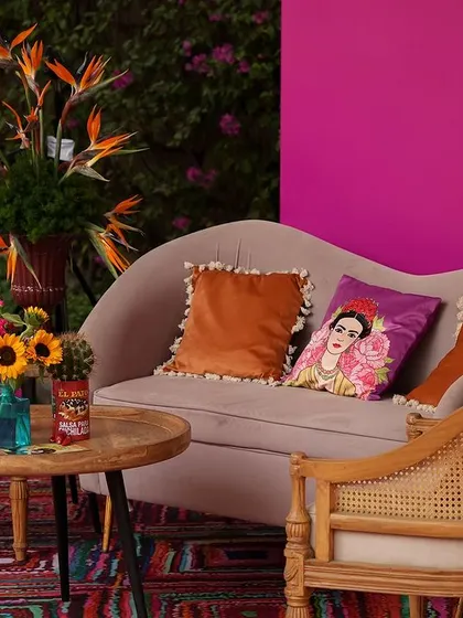 A close-up of a seating area, with a Frida Kahlo cushion and vibrant orange pillows. The background features a colorful, hand-painted mural.