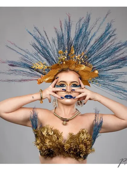 A creative beauty portrait featuring a dramatic headpiece made of gold and blue feathers. The bold makeup and direct gaze create a powerful, almost mythical, look.