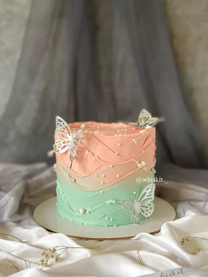 A beautiful ombre cake in peach and mint green, decorated with delicate silver butterflies and pearls. The wavy frosting adds a graceful touch.