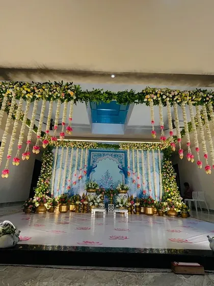 A beautiful stage setup with a Pichwai-inspired backdrop, framed by lush floral pillars and hanging jasmine garlands.