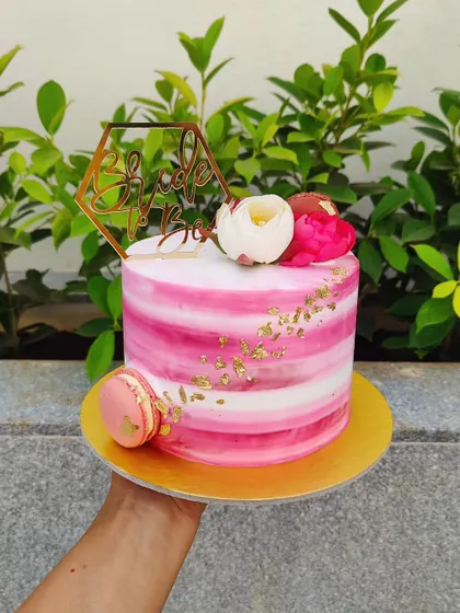 This bride-to-be cake features a pink and white watercolor stripe effect, accented with gold leaf, a macaron, and a custom topper.