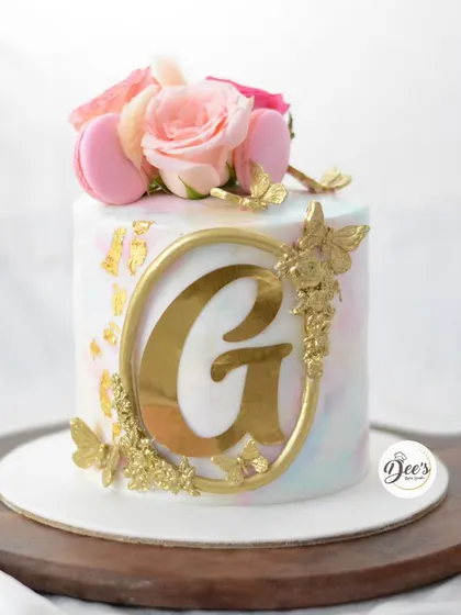 This monogram cake is a statement in elegance. A bold gold initial is surrounded by golden butterflies and topped with fresh roses and macarons, all against a soft watercolor background.