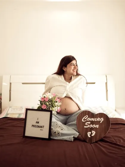 A joyful indoor portrait of the mom-to-be sitting on a bed, surrounded by flowers and "I am Pregnant" and "Coming Soon" signs.
