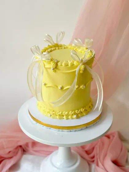 A bright and cheerful yellow cake with vintage-style piping and pearl details. The sheer white ribbons add a touch of delicate charm.