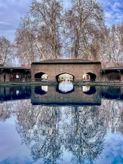 A perfect reflection of the Mughal arcade and bare trees in the crystal-clear water of Verinag spring. The stillness of the water creates a mirror to another world.