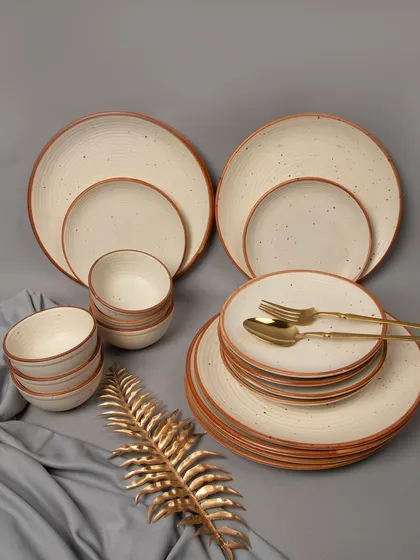 A complete cream-colored, speckled ceramic dinnerware set, elegantly arranged with a golden leaf prop and grey fabric.