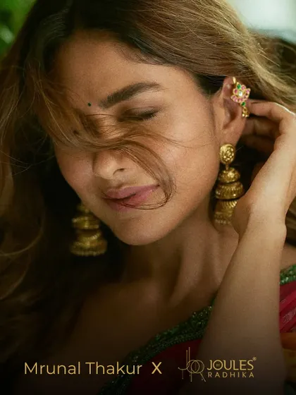 A beautiful, artistic shot of Mrunal Thakur, with her hair gently sweeping across her face, highlighting the intricate gold jhumkas and earcuff.
