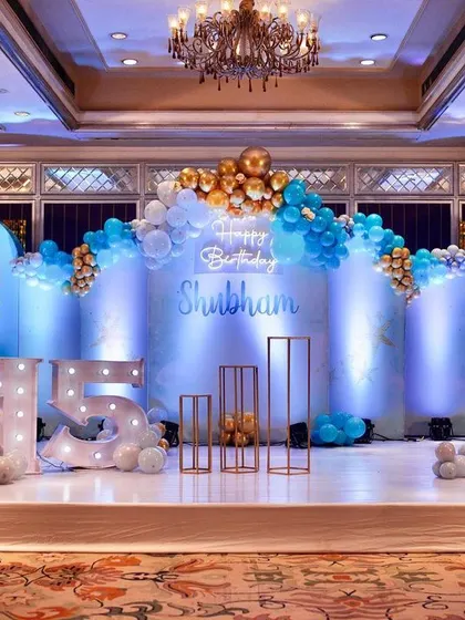 A cool and stylish 15th birthday party setup. I used a blue, white, and gold color palette with modern arched backdrops, balloon garlands, and light-up marquee numbers for a fun and elegant stage.
