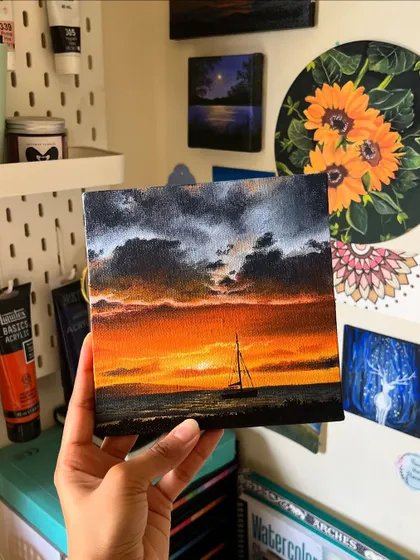 Holding up the finished painting of a sailboat at sunset. The dark clouds and fiery sky create a beautiful, dramatic contrast.