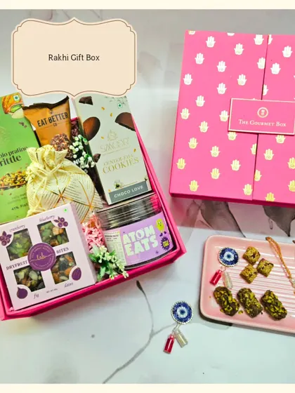 A beautiful shot of the pink Rakhi Gift Box, filled with treats like cookies, brittle, and dry fruit bites. It comes with a traditional Rakhi and a protective Evil Eye Roli Chawal set.