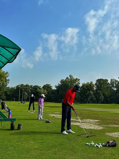 A golfer in a bright red shirt prepares to swing, with a junior golfer practicing nearby. It's a typical Sunday scene, showing the diverse community at our camp.