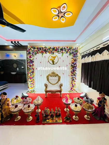 A wide view of a home-based baby shower. The floral backdrop, white throne, and traditional dolls create a complete and beautiful ceremonial space.