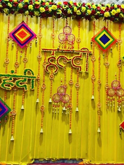 A combined 'Haldi Mehendi' backdrop in bright yellow, featuring floral signs for both functions and decorated with colorful hanging kites.