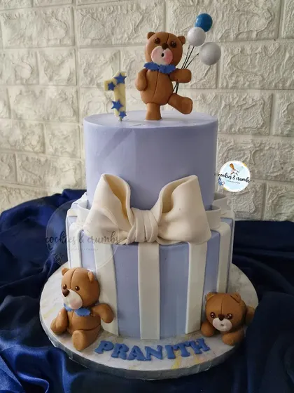 An adorable two-tiered teddy bear cake for a first birthday. The design includes a big fondant bow, stripes, and cute bears, creating a classic and sweet look for a little boy.