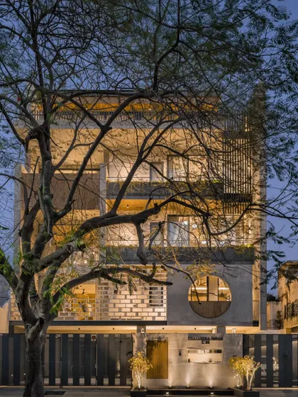 At night, the Swatantra Residence's façade is transformed by light. The illumination highlights the textural details of the concrete, the pattern of the jaali, and the warmth of the wood within the circular cutout, creating a dramatic street presence.