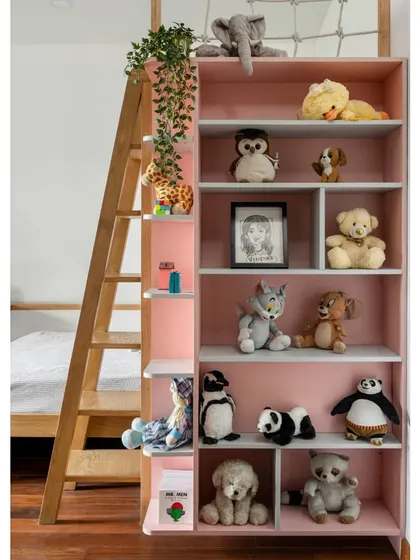 A close-up of the bookshelf in the 'Vasudhalayah' daughter's room, filled with stuffed animals and toys. The pink and white color scheme is playful and cheerful.