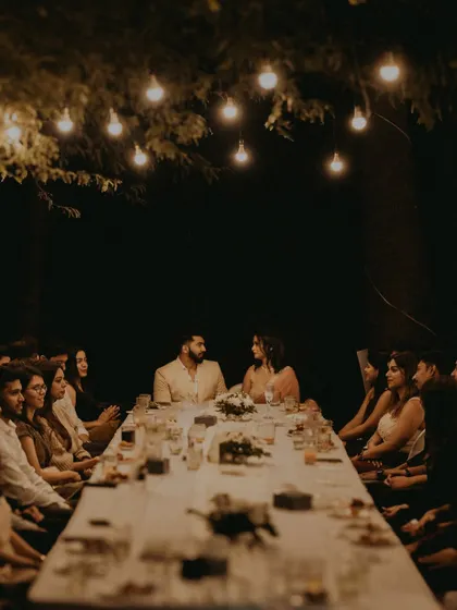 An atmospheric shot of the couple at their engagement dinner, seated at the head of a long table under hanging lights.