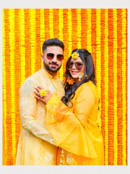 A classic couple portrait from a Haldi ceremony, set against a stunning wall of marigolds. The couple's coordinated yellow outfits and sunglasses add a touch of modern fun.