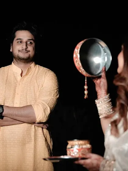 This image captures the traditional Karwa Chauth ritual with a focus on the husband's admiring gaze. The composition draws the viewer's eye to the interaction, telling a story of love and devotion.