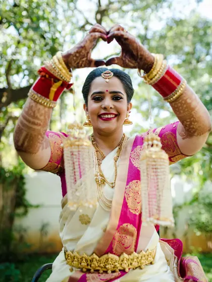 A joyful bride in traditional South Indian attire, making a heart shape with her hands. Her bright smile and the vibrant colors of her saree and kaleeras radiate pure happiness.