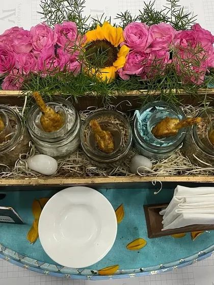 A rustic yet elegant presentation of various dips or small bites in glass jars. The arrangement on a bed of straw within a wooden crate, adorned with pink roses, creates a charming and memorable serving style.