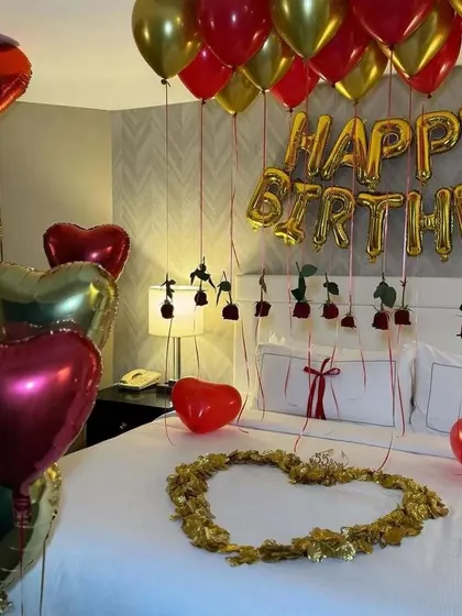 A classic romantic birthday bedroom decoration. The setup includes gold and red heart-shaped foil balloons, 'Happy Birthday' foil letters, and rose stems attached to the balloon strings.