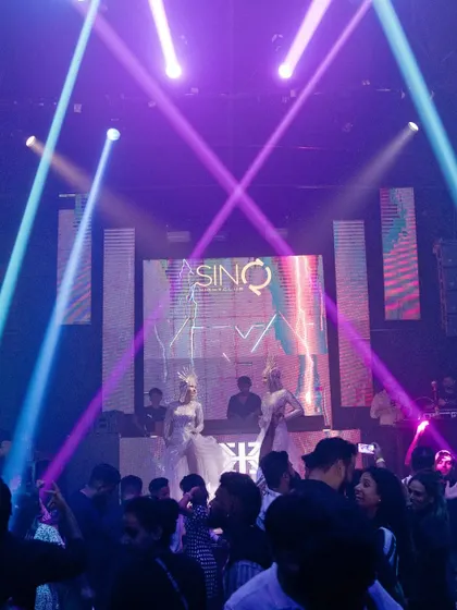 The stage at Sinq, lit with intersecting purple and blue beams. The visuals are a key part of the show, creating an unforgettable clubbing experience.