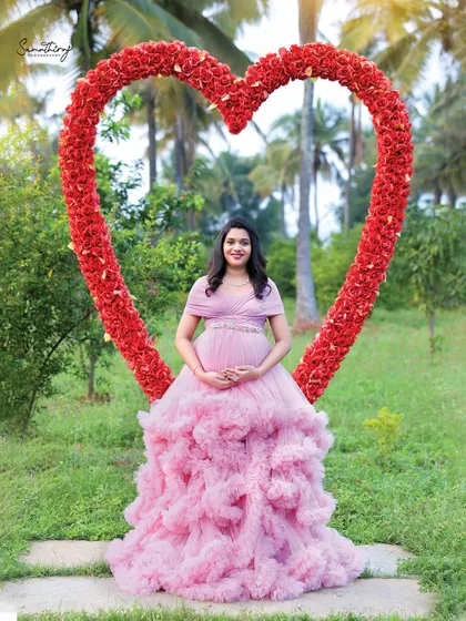 A solo portrait of a mother-to-be framed by a large heart-shaped floral prop. The pink ruffled gown and the romantic outdoor setting create a perfect fairytale moment.
