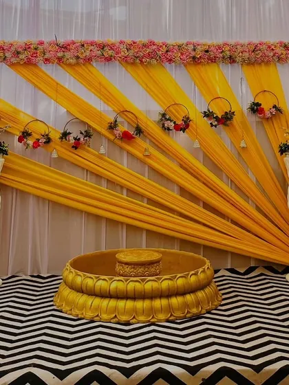 A simple yet elegant Haldi setup. The backdrop uses yellow drapes arranged in a sun-ray pattern, framed by a delicate floral border. The patterned rug adds a modern touch to the otherwise traditional decor.